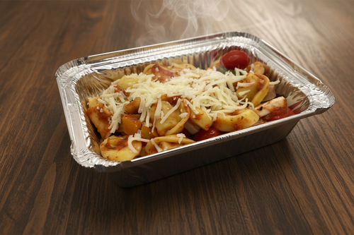 A foil container of baked tortellini with cheese on top, alongside vegetables and sausage visible beneath the surface.